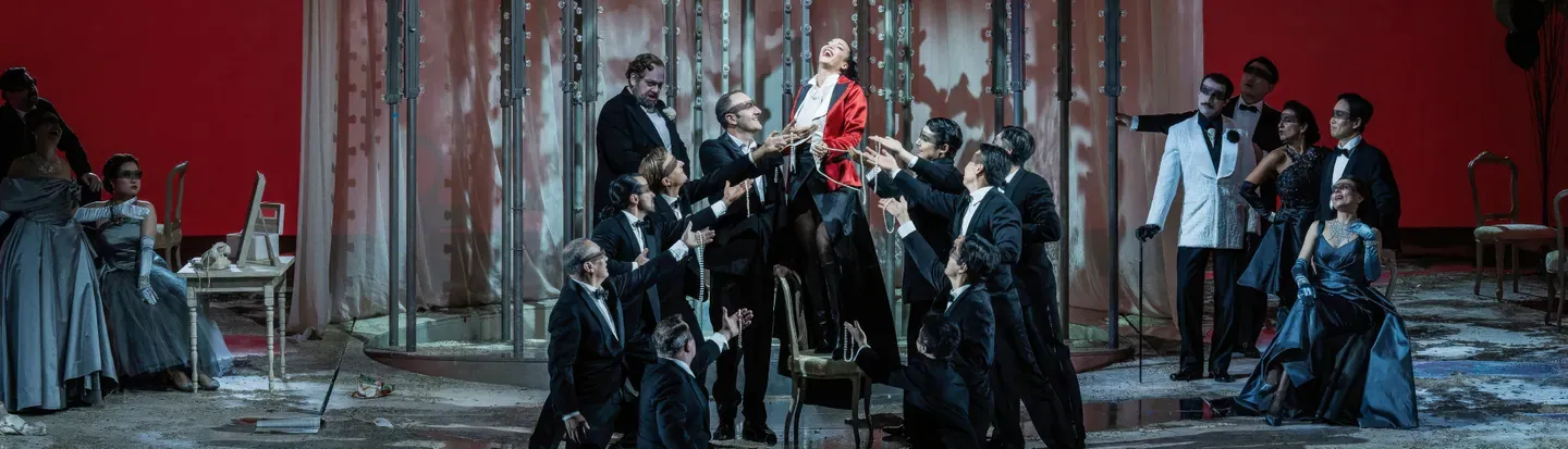 Escena de Manon Lescaut con la protagonista rodeada por otros personajes ante un telón rojo